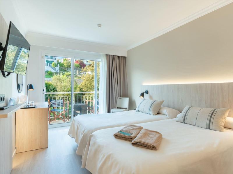 Helles Hotelzimmer mit zwei Betten, Fernseher und Balkon mit Blick auf Grünflächen.