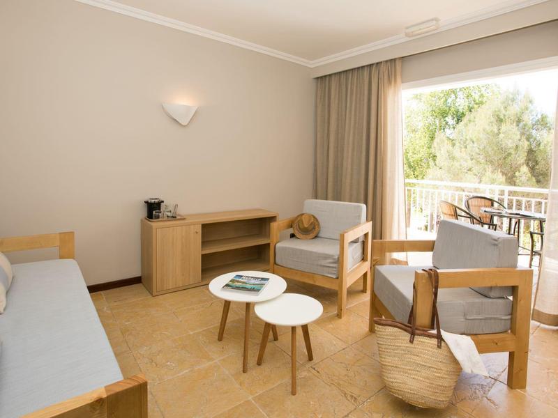 Helles Wohnzimmer mit Sofa, Sesseln, Couchtischen und Balkon mit Blick auf grüne Bäume.