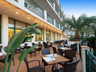 Area esterna di un hotel con terrazza coperta, tavoli in legno e sedie nere al crepuscolo.