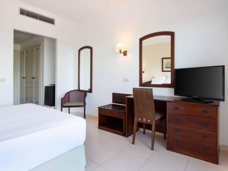 Camera d'hotel con letto, mobili in legno, specchio e TV contro parete bianca.