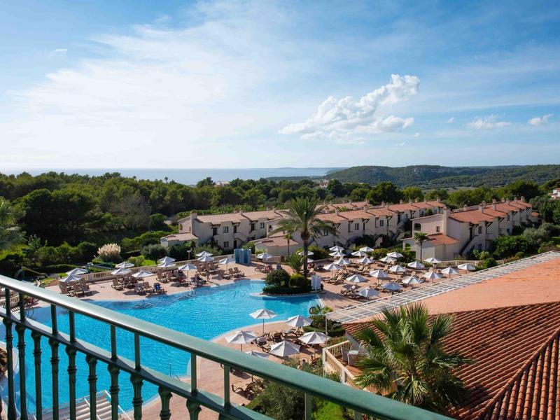 Vista di un resort con piscina, ombrelloni e edifici sotto un cielo azzurro.