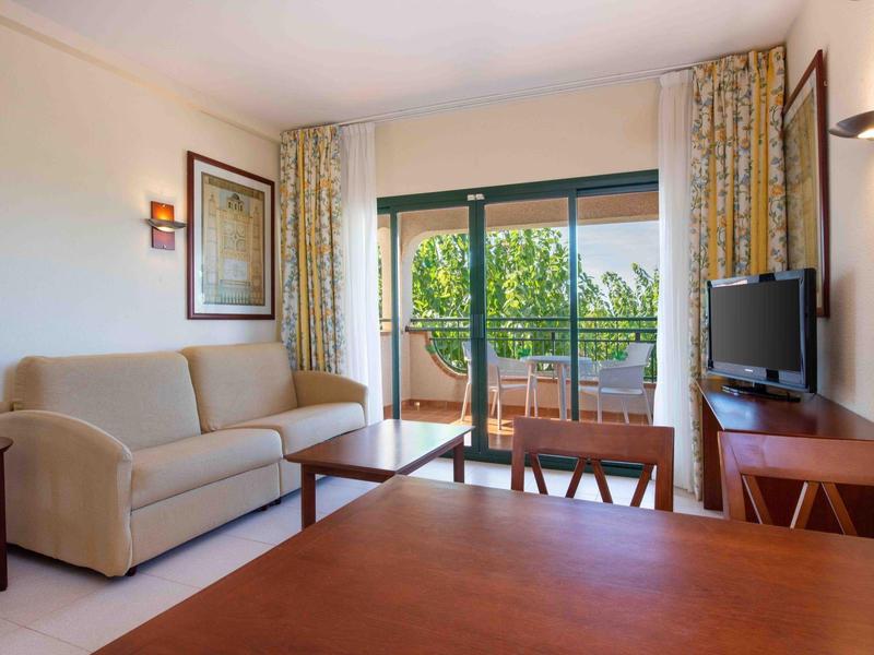 Camera d'hotel accogliente con divano, tavolo, TV e balcone con vista sul verde.