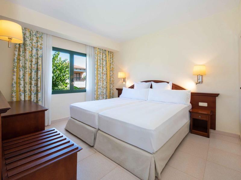 Camera d'albergo luminosa con due letti, comodini, lampada e finestra con tende.