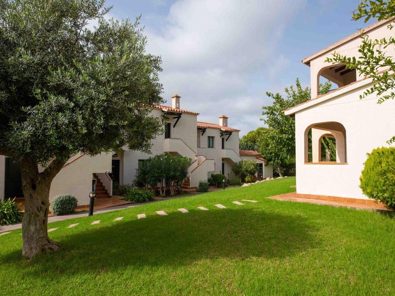 Area giardino verde con grandi alberi e case vacanze bianche sotto un cielo nuvoloso.