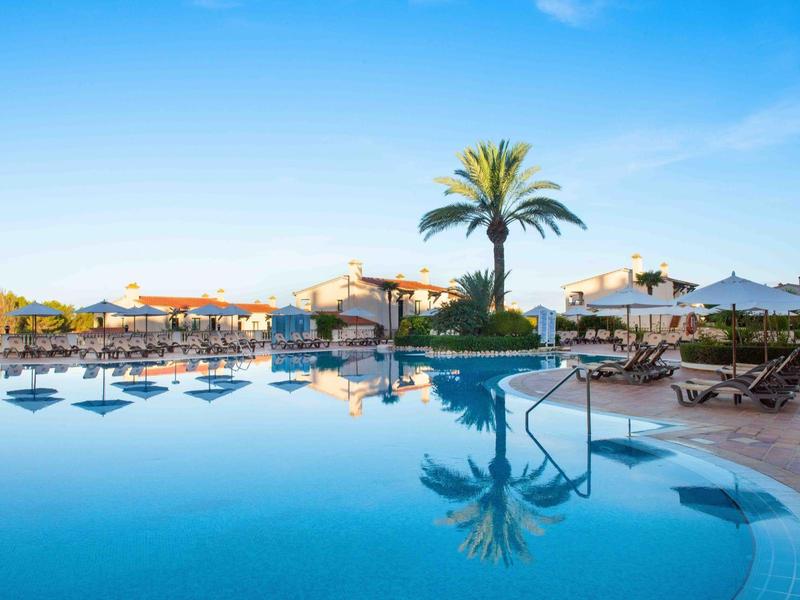 Piscina rotonda dell'hotel con lettini, ombrelloni e palme sotto un cielo azzurro