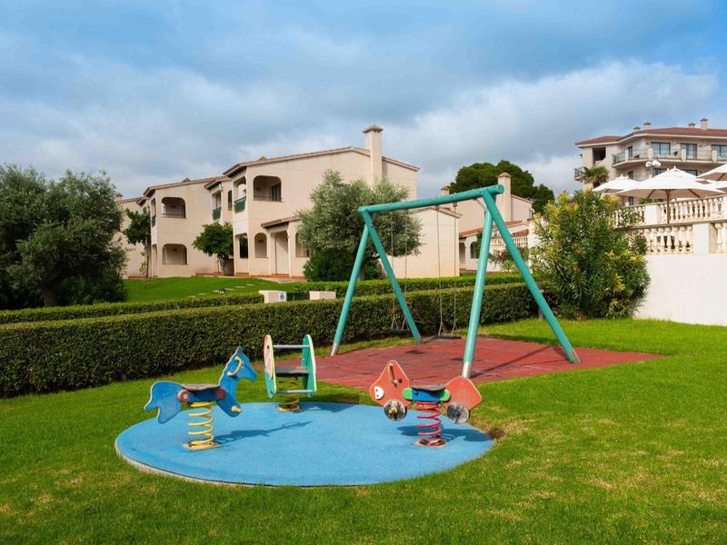 Parco giochi per bambini con altalene e seesaw su prato verde vicino a edifici residenziali.