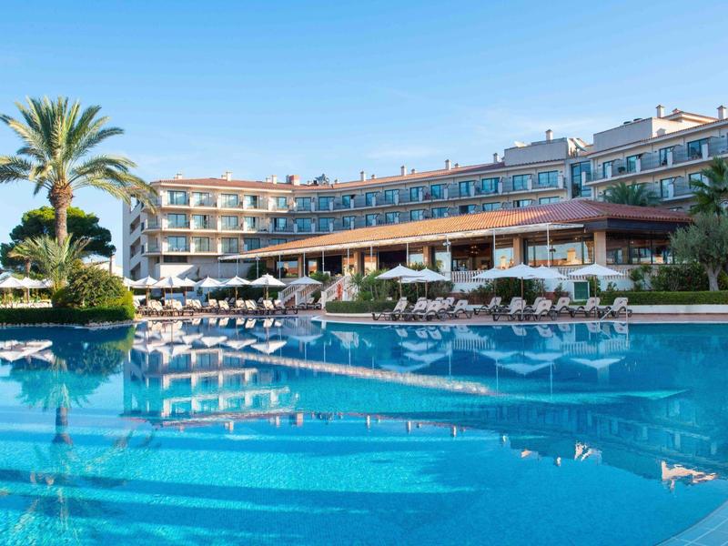 Grande edificio dell'hotel con piscina all'aperto, ombrelloni e palme sotto un cielo sereno.