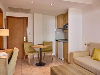 Przytulny pokój hotelowy z sofami, stołem, krzesłami i małą kuchnią w ciepłych kolorach.