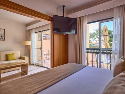 Jasny pokój hotelowy z podwójnym łóżkiem, sofą, telewizorem i balkonem z widokiem na zielone drzewa.