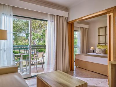 Jasny pokój hotelowy z balkonem, sofą i łóżkiem podwójnym, z widokiem na zielone drzewa.