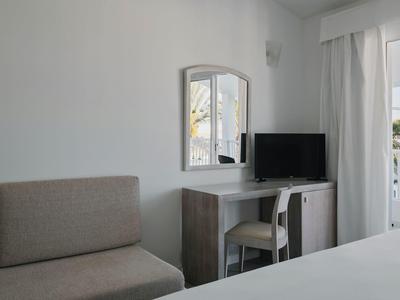 Eenvoudige hotelkamer met bank, bureau, stoel en flatscreen-tv naast balkon met gordijnen.