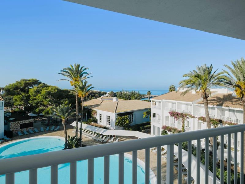 Uitzicht op een hotelzwembad met palmbomen en zee op de achtergrond bij heldere lucht.