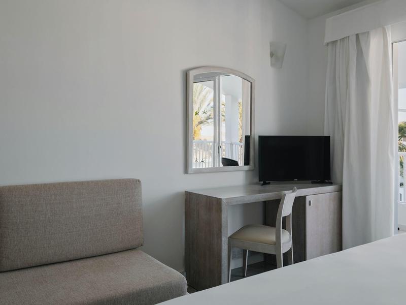 Eenvoudige hotelkamer met bank, bureau, stoel en flatscreen-tv naast balkon met gordijnen.