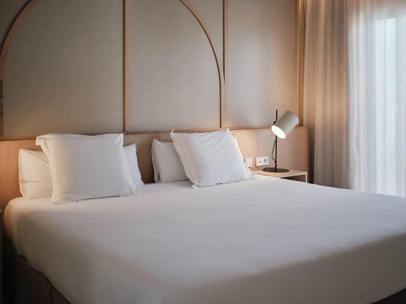 Modern ingerichte hotelkamer met groot bed, witte kussens en daglicht door gordijn.