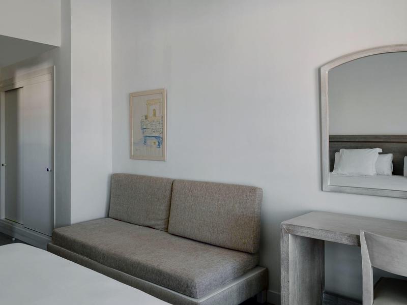 Een eenvoudig ingericht hotelkamer met een grijze bank, spiegel en nachtkastje in neutrale kleuren.