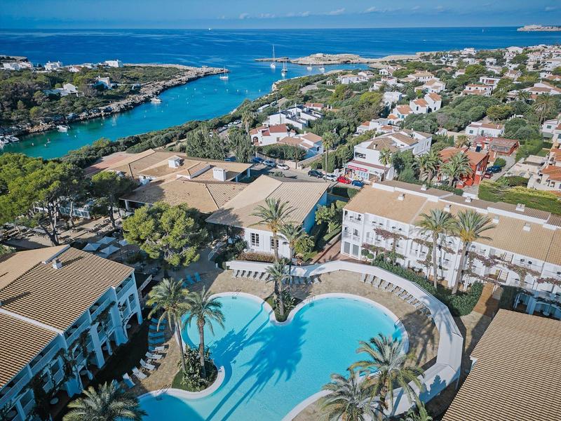 Hotel met groot zwembad en veel kamers vlakbij de kust en woonhuizen op de achtergrond.