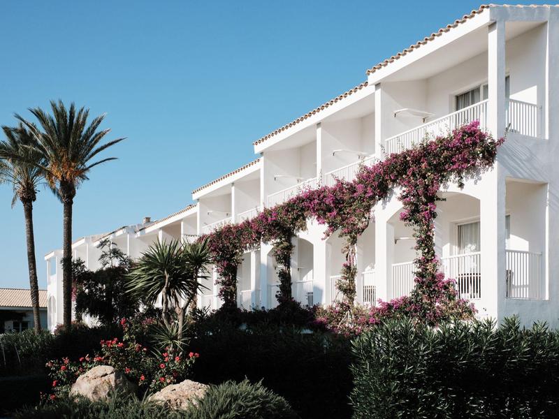 Wit twee verdiepingen tellend hotel met bloeiende klimplanten en palmbomen onder een heldere blauwe lucht.