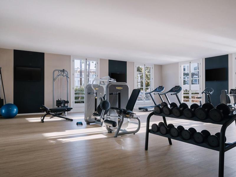 Lichte sportschool met diverse fitnessapparatuur en halters bij grote ramen.