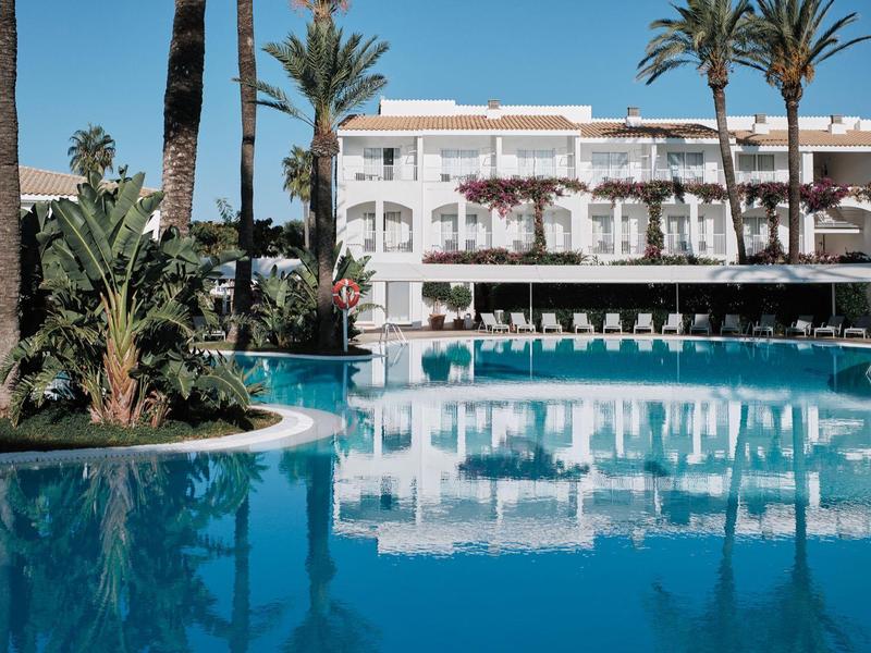 Groot buitenzwembad voor een wit meerlagig hotelgebouw met palmbomen.