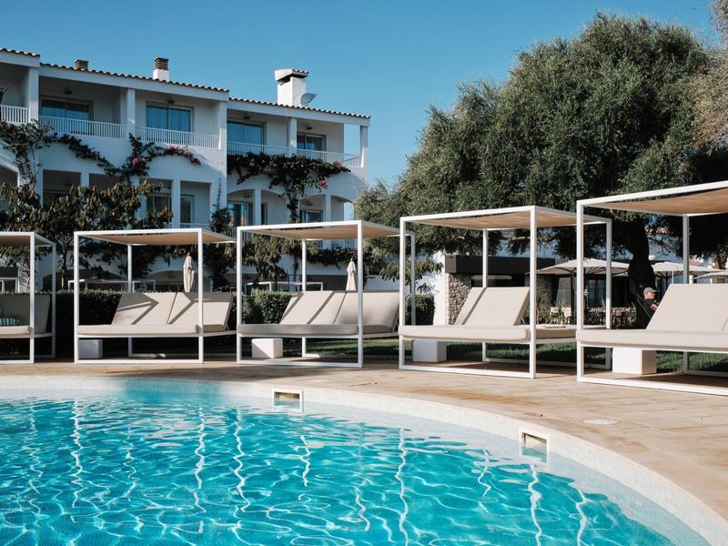 Comfortabele ligbedden met overkapping naast een blauw zwembad voor een wit hotel en bomen.