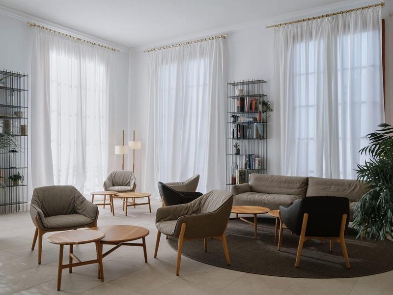 Lichte woonkamer met moderne fauteuils, bank, houten meubels en grote ramen met witte gordijnen.