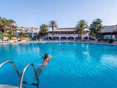 Large outdoor pool with a person swimming and surrounding palm trees and buildings.