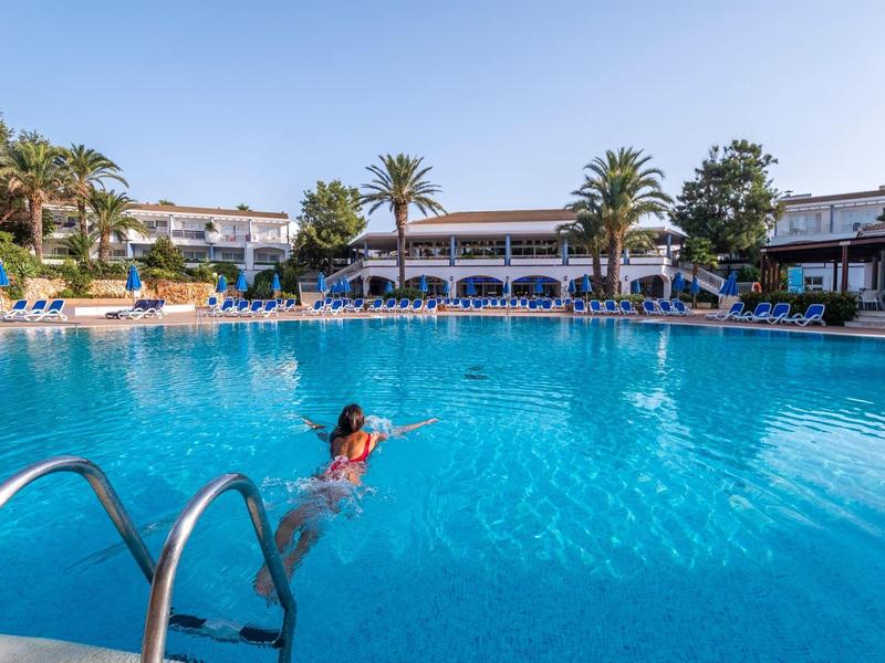 Large outdoor pool with a person swimming and surrounding palm trees and buildings.