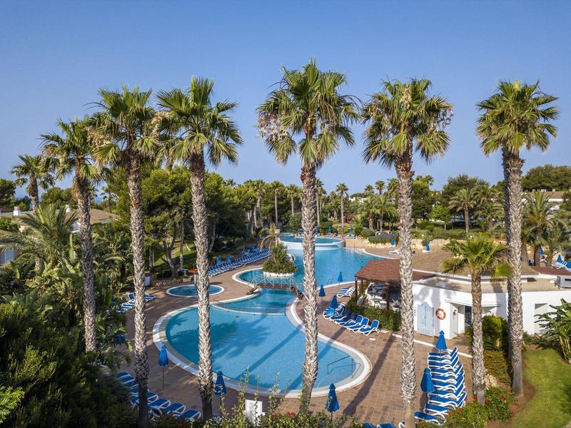 Hotel pool with palm trees and blue sun umbrellas on a sunny day.