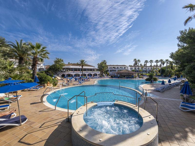 Large pool area with whirlpools, sun loungers, and palm trees under blue sky.