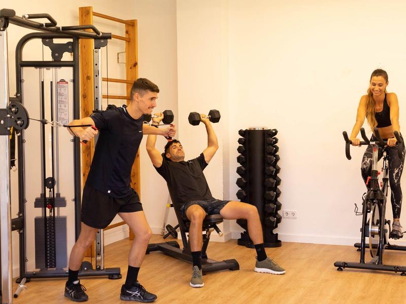 Three people exercise in a gym with dumbbells, a stationary bike, and strength equipment.