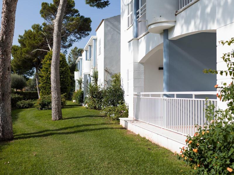 White hotel buildings with balconies next to a well-kept lawn and trees under a clear sky.
