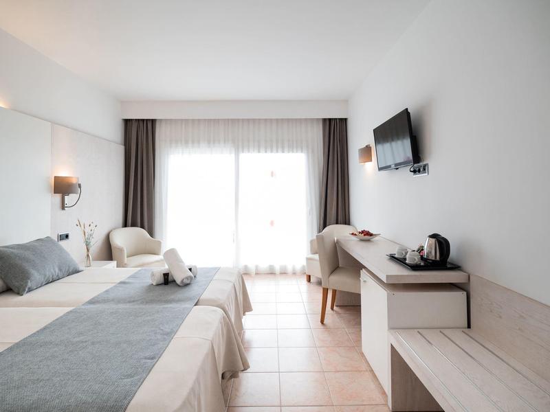 Bright hotel room with two beds, desk, TV, and balcony with curtains.