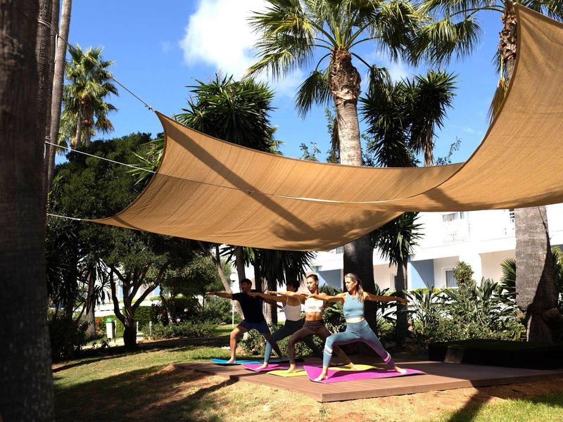 Two people practice yoga under sunshades in a garden with palm trees.