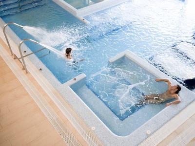 Two people relaxing in a modern indoor pool with jets and clear water, wooden floor nearby.