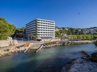 Modern hotel beside clear coastal water with greenery and blue sky in the background.