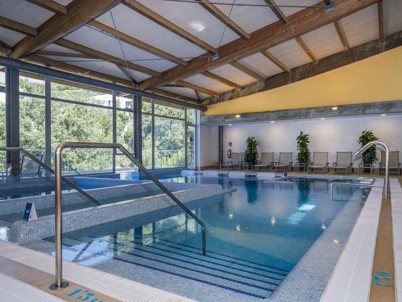 Indoor hotel pool with large windows, plants, and lounge chairs under a wooden roof.