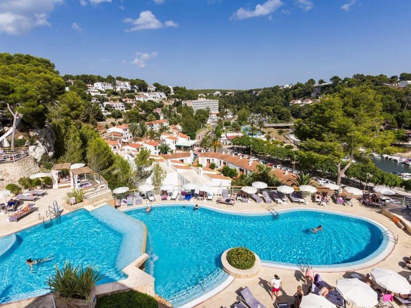 A large outdoor hotel pool with sun loungers and umbrellas surrounded by trees and buildings.