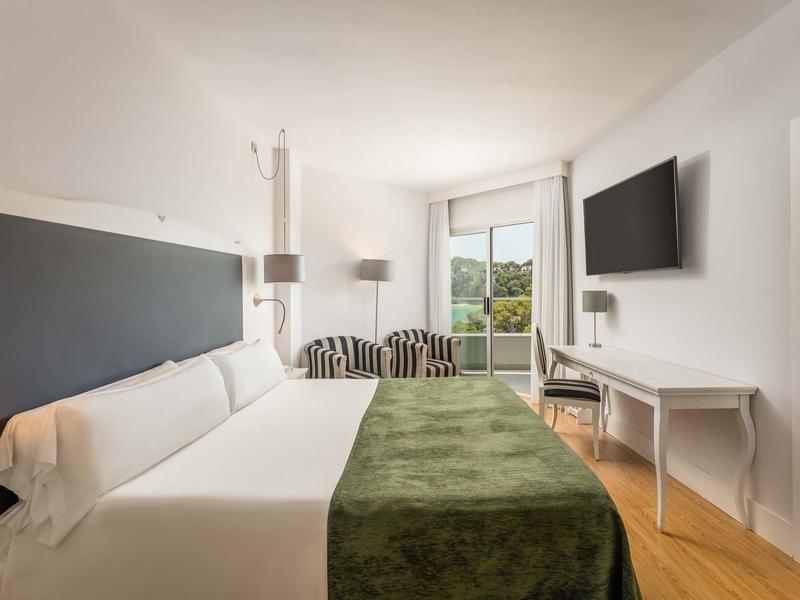 Bright hotel room with king bed, TV, desk, and balcony with green view.