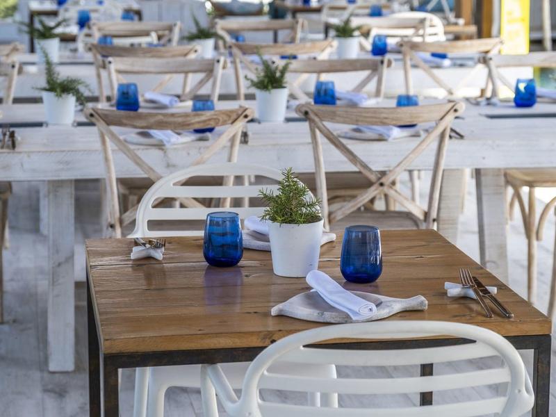 Outdoor restaurant seating with wooden tables, white chairs, blue glasses, and potted plants.