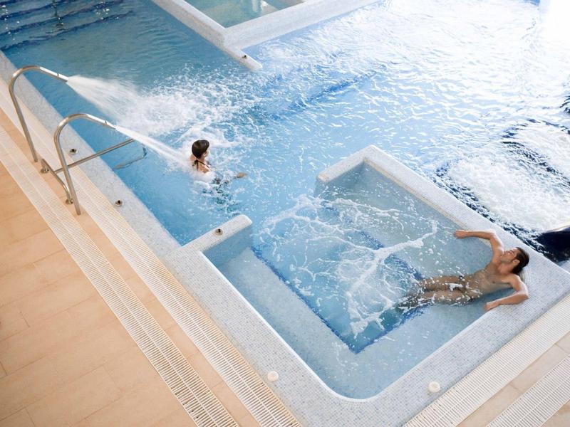 Two people relaxing in a modern indoor pool with jets and clear water, wooden floor nearby.