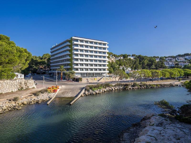 Modern hotel beside clear coastal water with greenery and blue sky in the background.