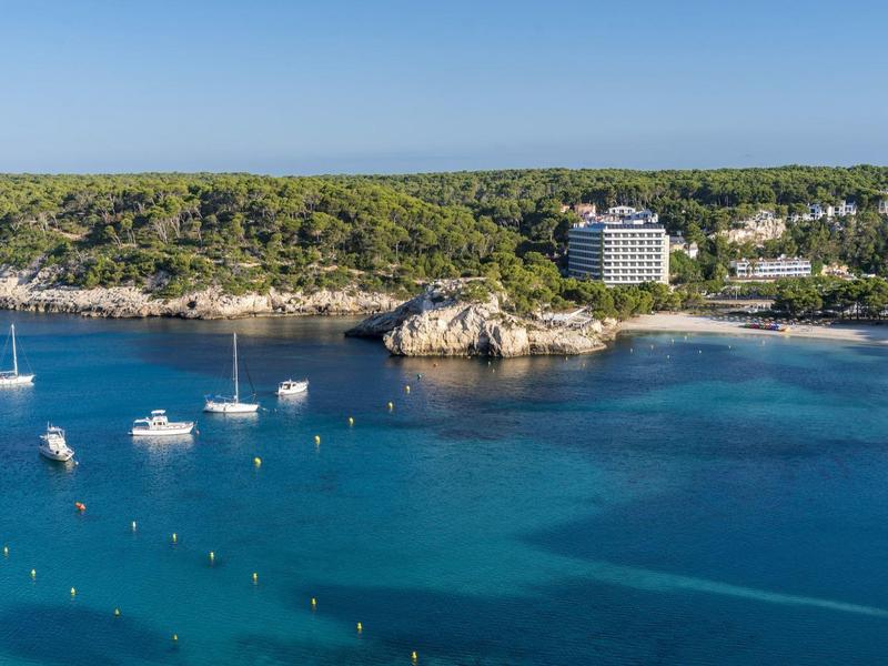 Coastal hotel with boats on clear blue water and green forested hills under a clear sky.