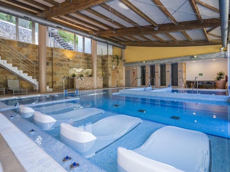 Indoor pool with lounge chairs submerged in water under wooden ceiling and stone walls.