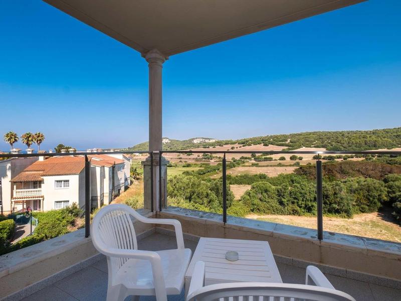 Balcon avec chaises en plastique et table, vue sur des maisons et des collines verdoyantes sous un ciel bleu.