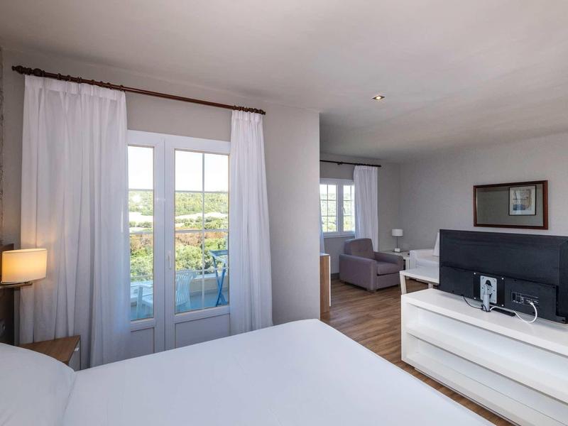 Chambre d'hôtel lumineuse avec lit double, télévision sur commode et terrasse donnant sur la verdure.