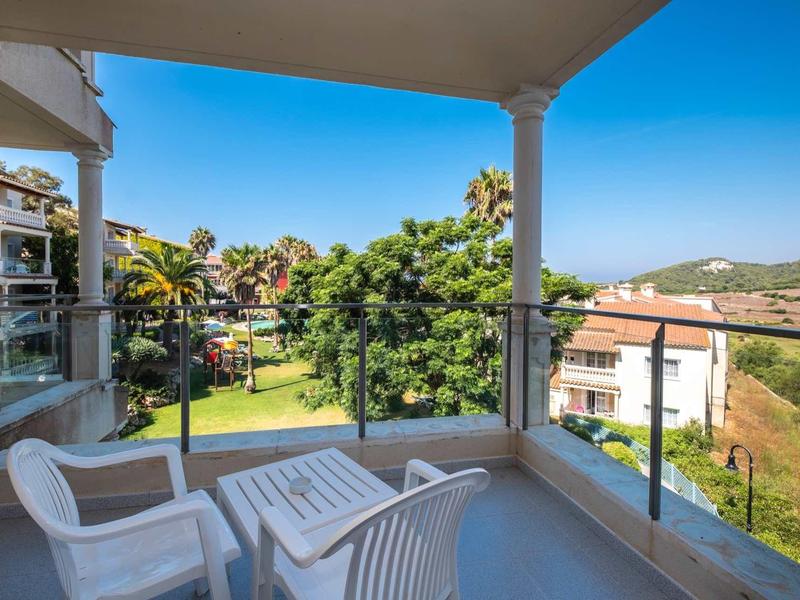 Balcon avec chaises blanches et table donnant sur des jardins verts et des bâtiments.
