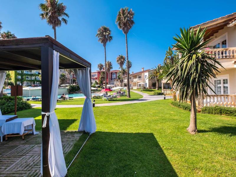 Jardin vert avec gazebo et palmiers devant des bâtiments d'hôtel sous un ciel bleu.