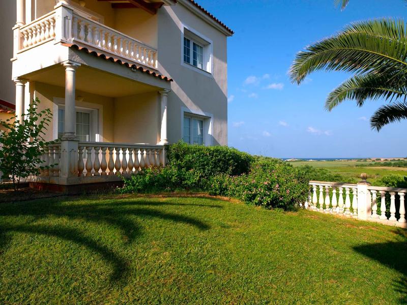 Jardin d'hôtel ensoleillé avec pelouse, balcon et palmiers sous un ciel bleu clair.