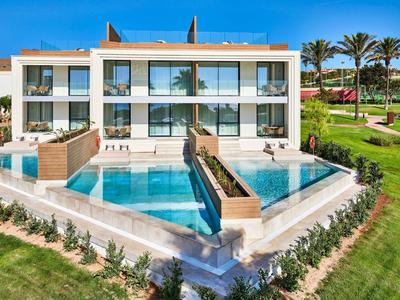 Modern hotel building with two pools and manicured garden under a blue sky.