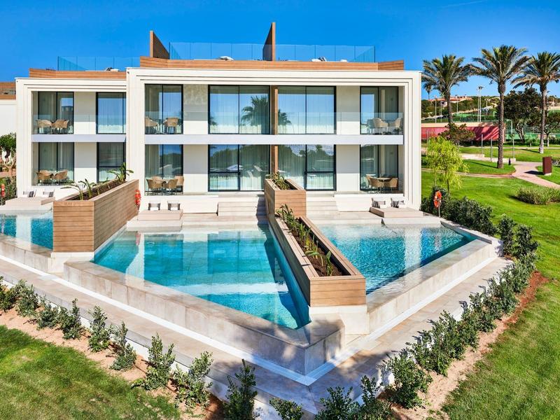 Modern hotel building with two pools and manicured garden under a blue sky.